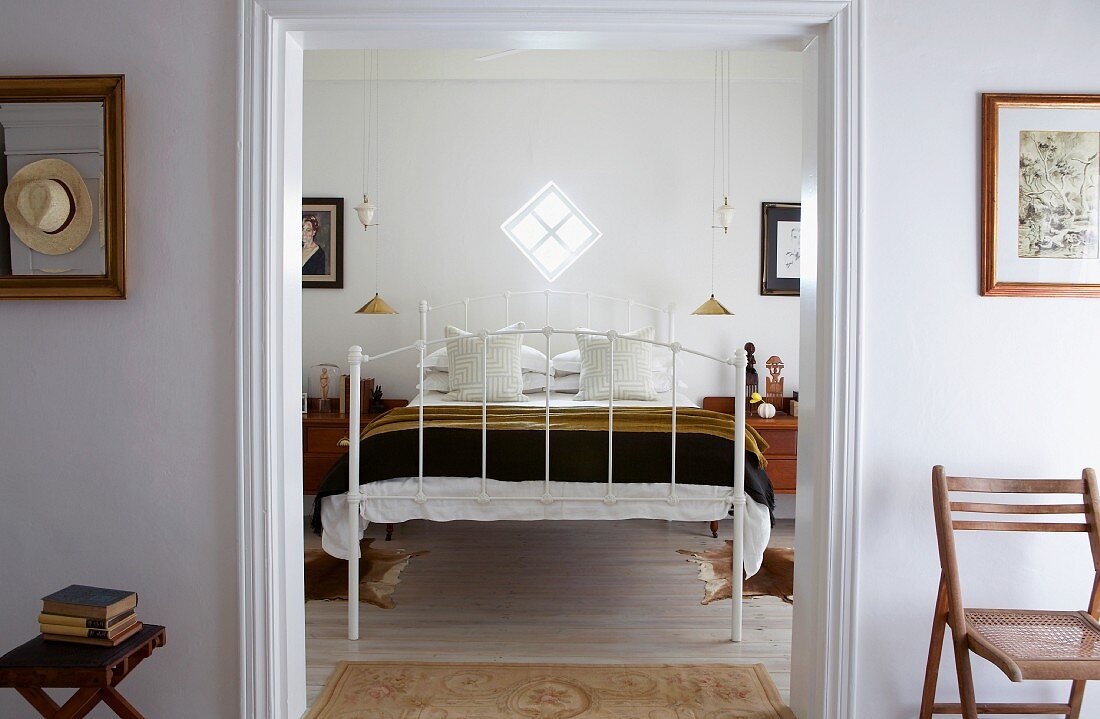 White metal bed below small, lozenge-shaped window; gilt pendant lamps flanking bed