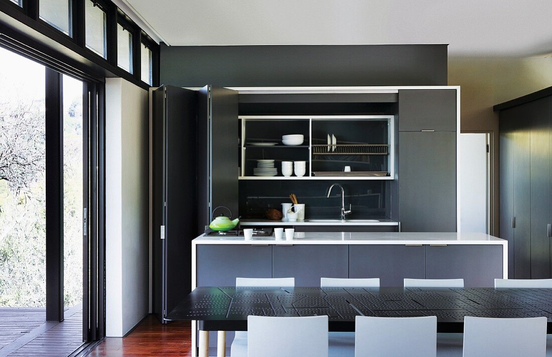 Einbauküche mit Falttüren und Küchenblock hinter Esstisch mit schwarzer Metallplatte; seitlich offene Schiebetüren zur Terrasse