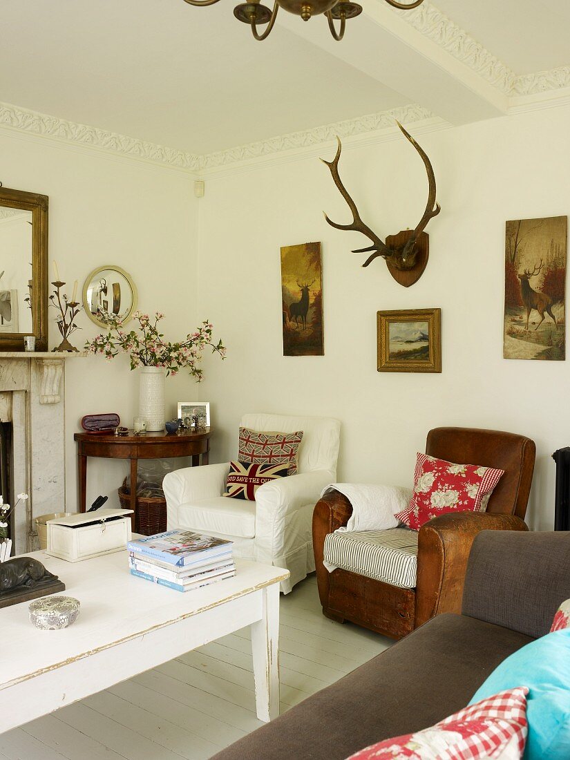 Scatter cushions on armchairs, white-painted wooden table, sofa and pictures with hunting motifs and antlers in living room