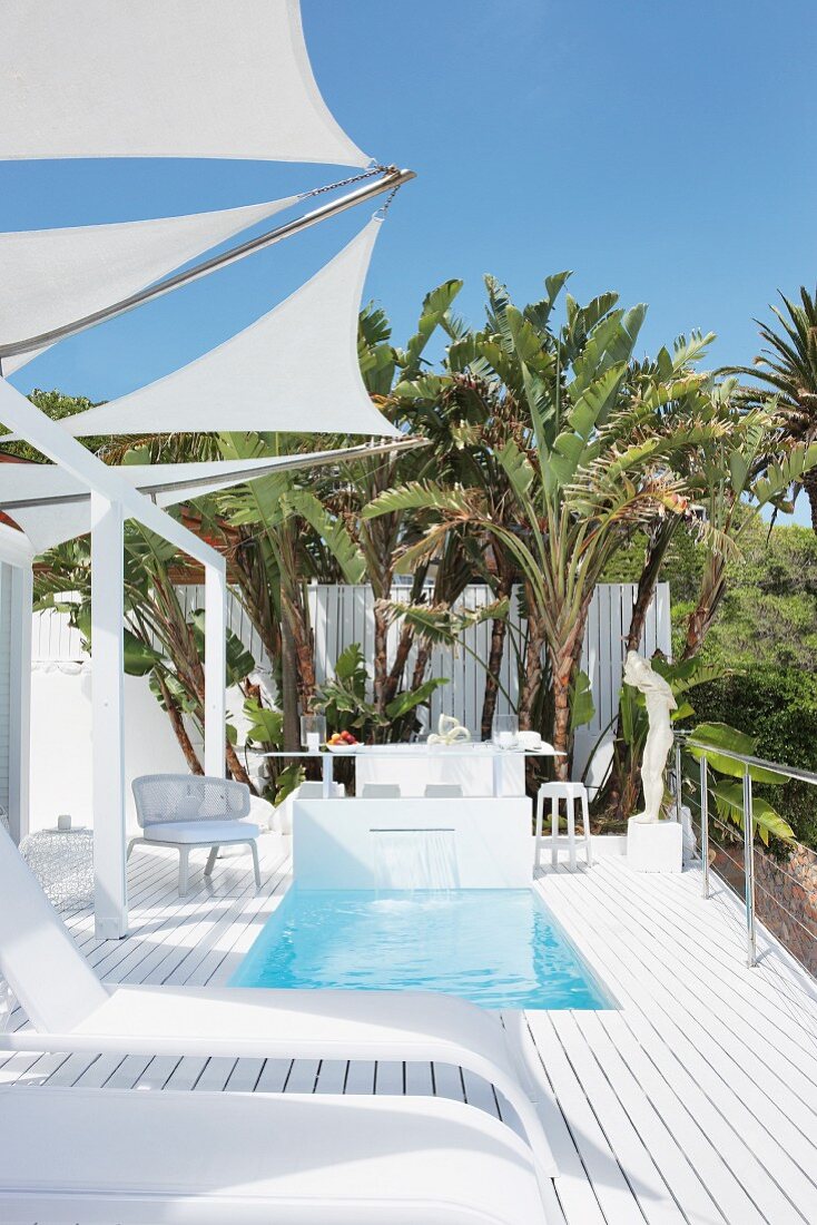 Designer Sonnenliegen auf weisser Terrasse unter dreieckigen Sonnensegeln und eingelassenem Pool vor Palmen