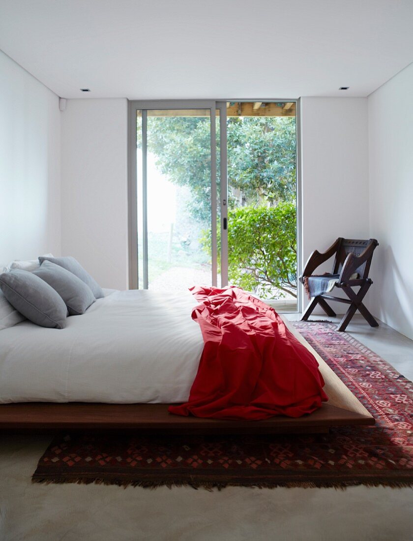 Schlichtes Doppelbett mit roter Tagesdecke auf Orientteppich vor Schiebe-Terrassentür und Blick in Garten