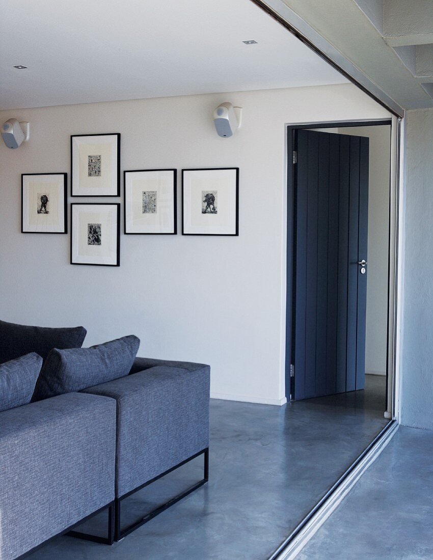 Offene, blaue Holztür mit Wohnzimmercouch im selben Farbton und einer Sammlung Tuschezeichnungen an der Wand vom Balkon aus gesehen