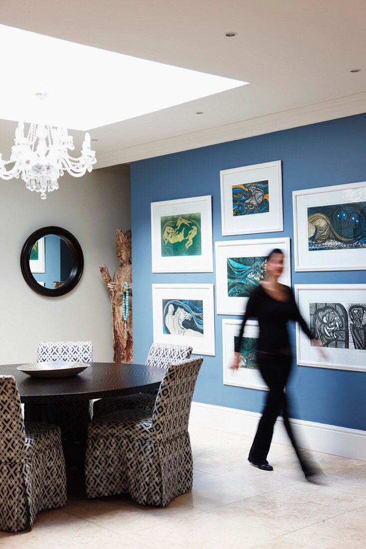 Moderne Kunst an blauer Wand; unter einem großem Lichtfenster ein runder Esstisch und Stühle mit gemusterten Hussen
