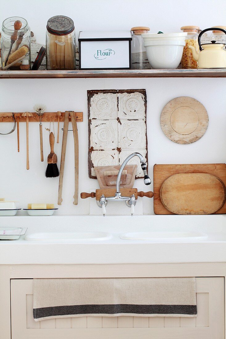 Kitchen sink below kitchen utensils hanging on wall and various vessels on shelf