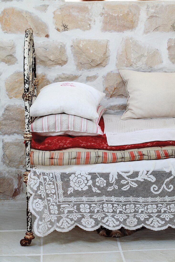 Spitzendecke unter Kissen- und Deckenstapel auf Tagesbett mit verrostetem Metallgestell vor Natursteinwand