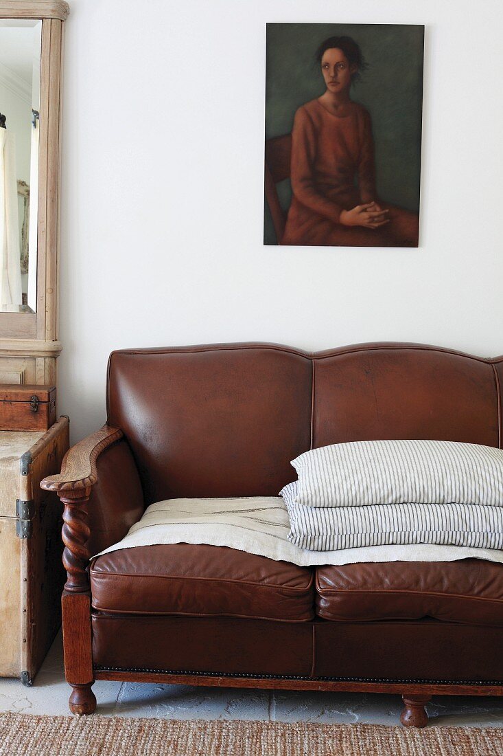 Stacked scatter cushions on traditional, brown leather sofa below painting of woman on white wall