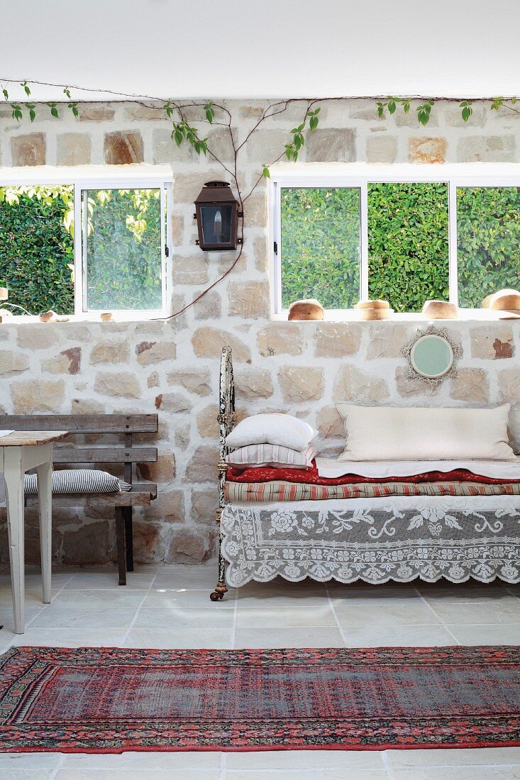 Traditioneller Teppichläufer auf Boden vor Vintage Tagesbett mit Kissen unter Fenster an Natursteinwand