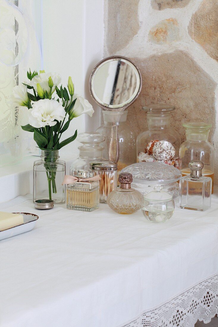Schlichter Tisch mit weisser Spitzendecke und einer Sammlung von Vintage Flacons neben Blumenstrauss und Handspiegel vor Natursteinwand