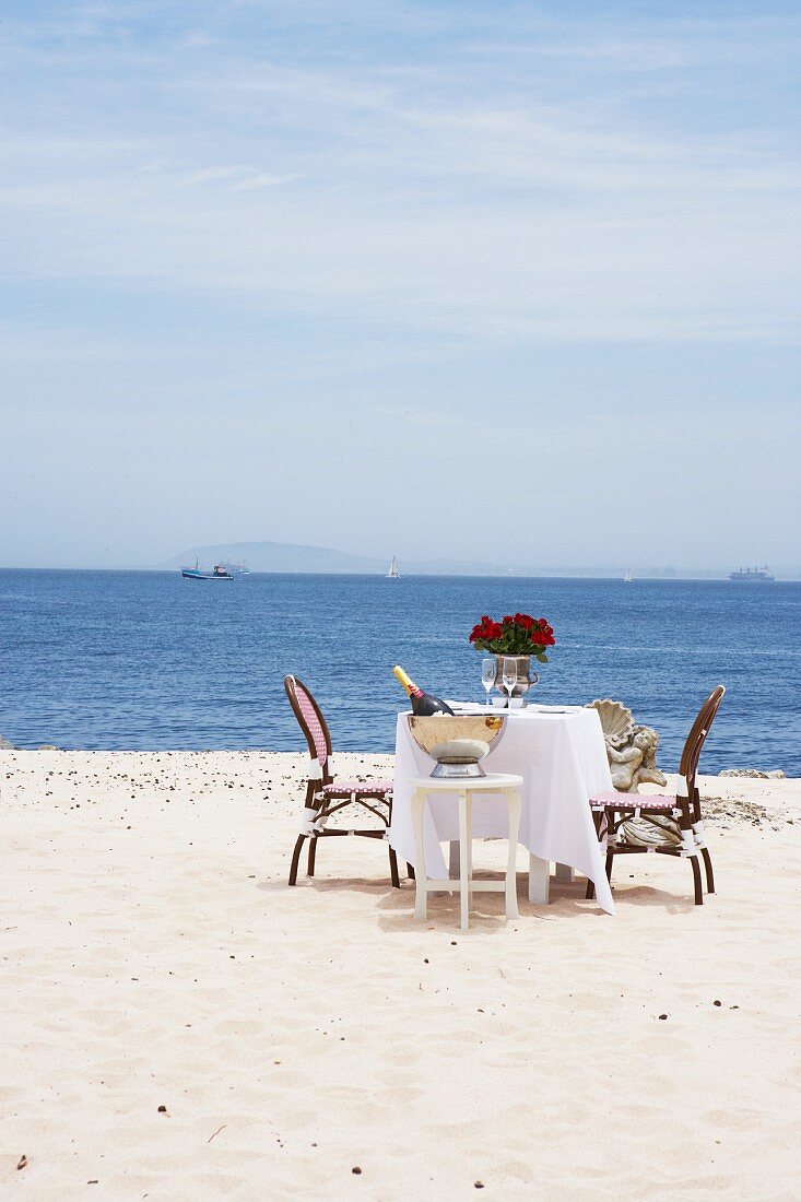 Gedeckter Tisch für Zwei mit Sekt und Blumen am Sandstrand am Meer