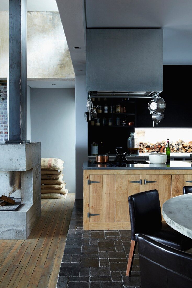 Open-plan interior with black brick floor and wooden kitchen cupboards; concrete open fireplace to one side
