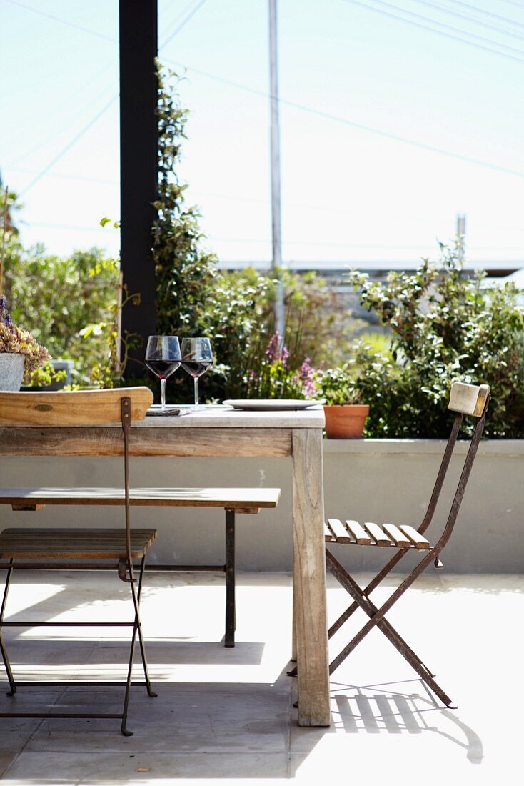 Sonnige Terrasse mit Vintage Tisch und rostigen Klappstühlen; auf dem Tisch Teller und zwei Rotweingläser