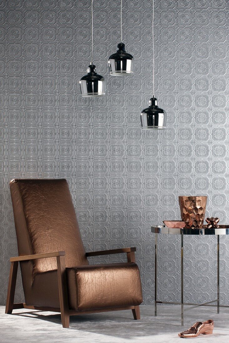 Reading chair with metallic brown cover against silver wallpaper and below three chrome pendant lamps