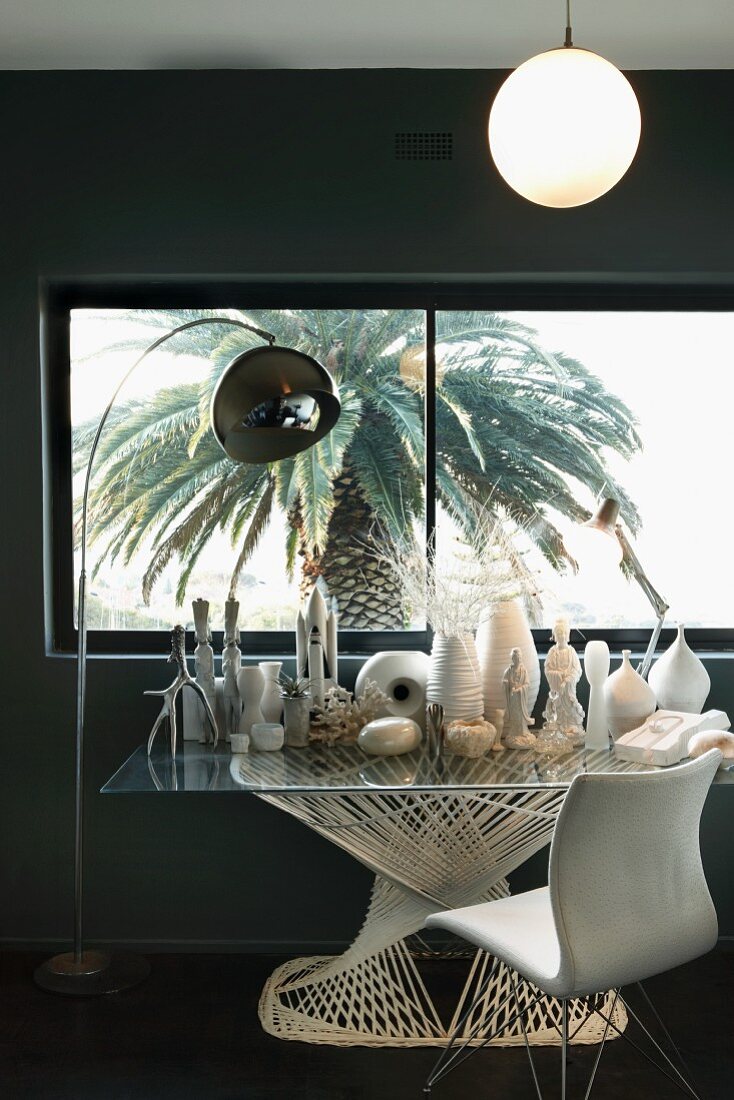 Sammlung aus weissen Vasen und Skulpturen auf modernen Glastisch neben Bogenlampe und Vintage Stuhl vor Fenster mit Blick auf Palme