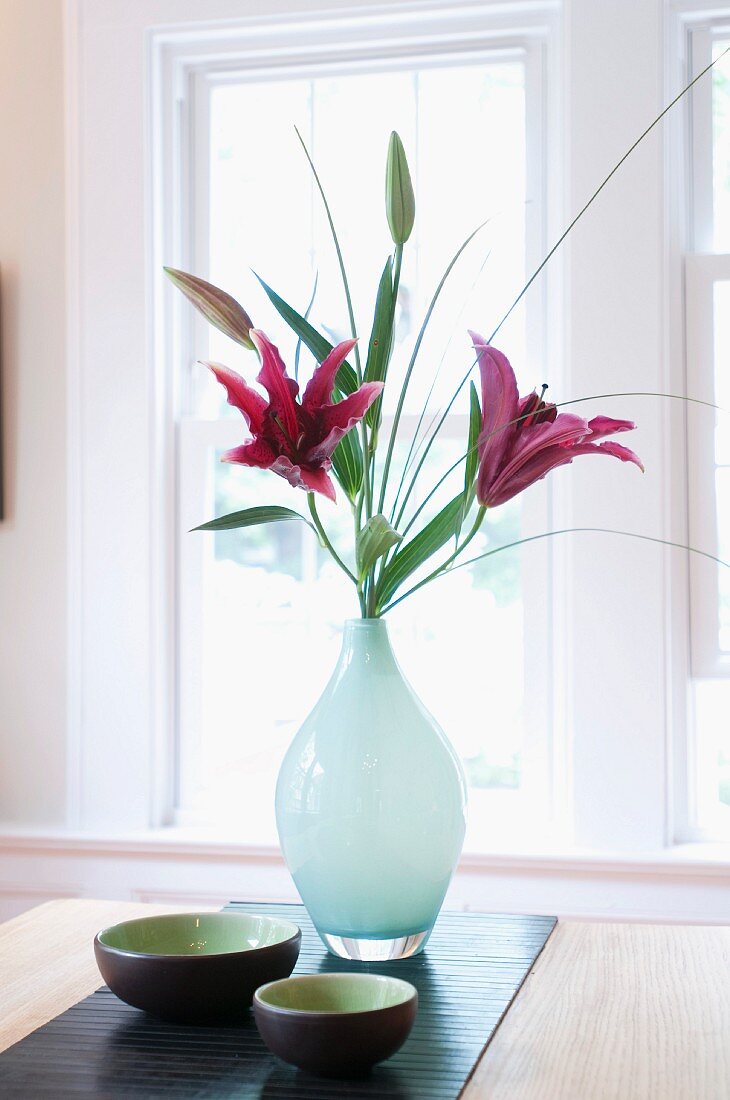 Pink lilies in glass vase and Japanese dishes on bamboo runner in front of window