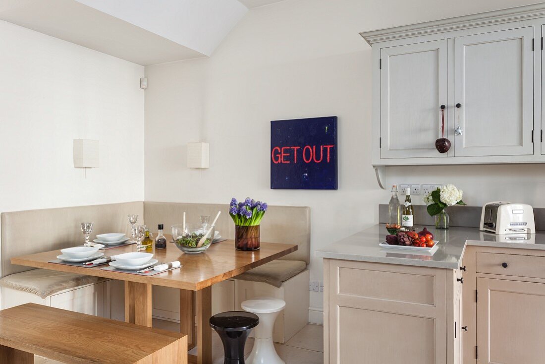 Blaurotes Schriftbild hinter quadratischem Esstisch mit Eckbank und schlichte Küche im dezent farbigen Shakerstil