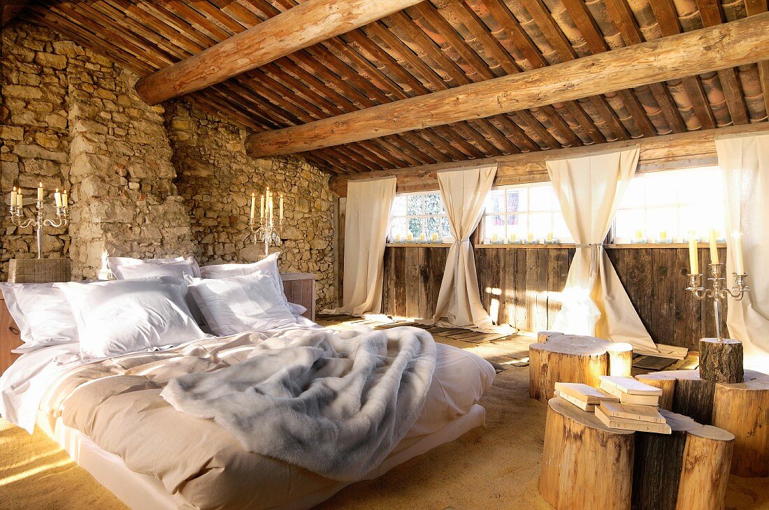 Double bed and tables made of tree trunks on sand floor in barn-like interior with rough stone walls and gathered curtains at window