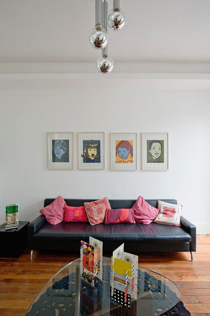 Glass coffee table opposite black leather couch against wall below Warhol screen prints in minimalist interior