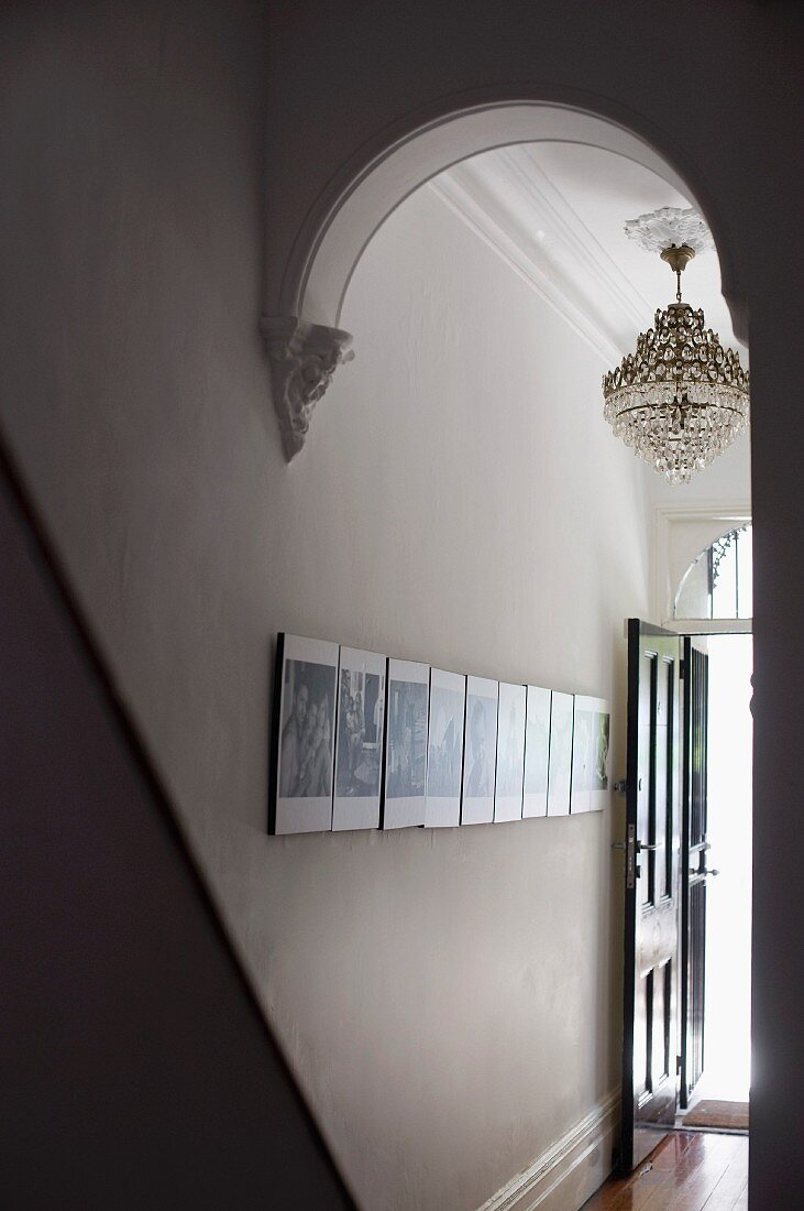 View through arched doorway of chandelier and gallery of photos on wall of narrow hallway of grand apartment