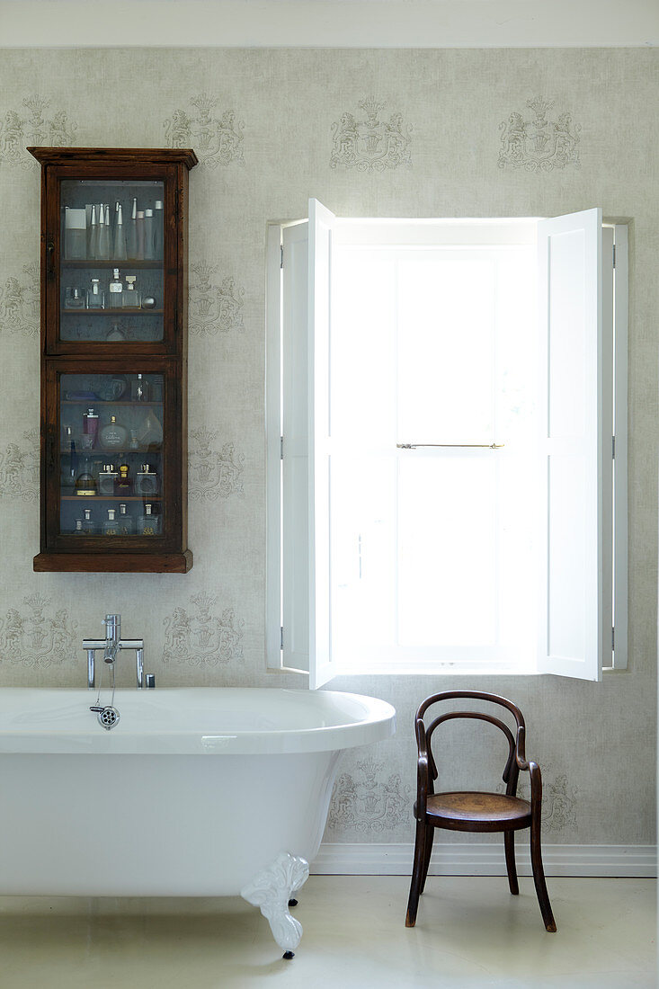Traditional bathroom with glass-fronted wall cabinet above retro bathtub and classic bentwood chair; window with interior shutters