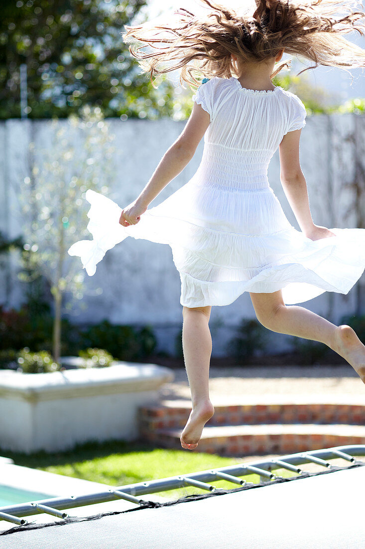 Mädchen mit romatischem, weißem Sommerkleid springend auf Trampolin im Garten