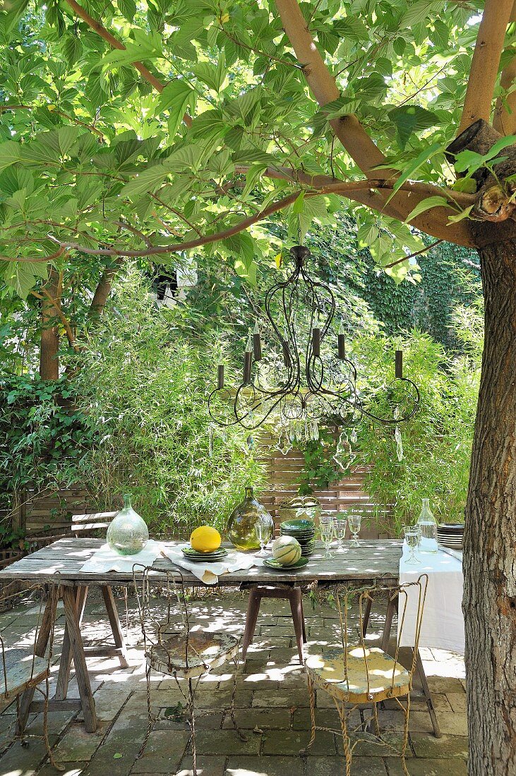 Rustic table and vintage metal chairs under candle chandelier hanging from tree in Mediterranean garden
