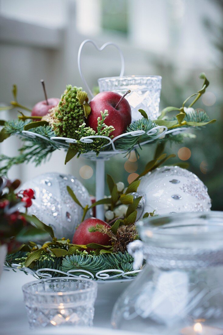 Weihnachtsgesteck auf einer Etagere mit Äpfeln, Tannenzweigen, Misteln, Moos, Windlicht und Christbaumkugeln
