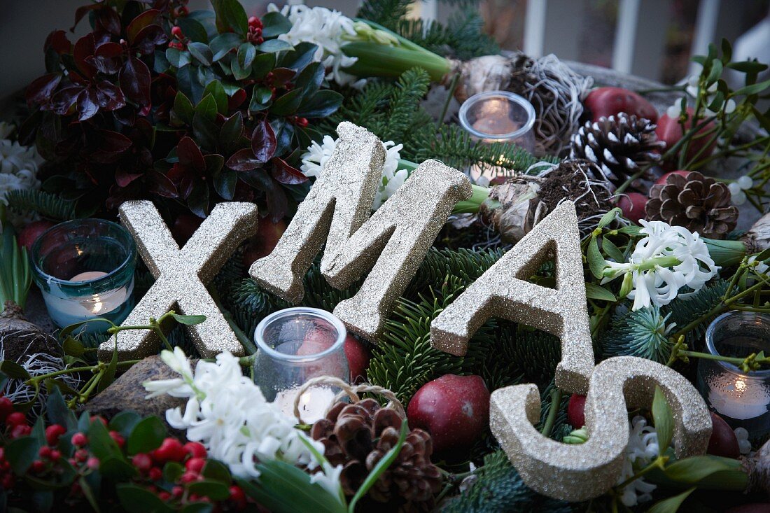 Weihnachtsgesteck aus Tannenzweigen, Scheinbeeren, Lorbeer, Äpfeln, Misteln, Hyazinthen und Pinienzapfen mit Windlicht und Buchstaben XMAS