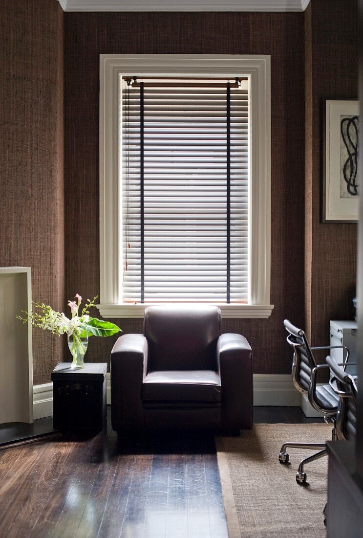 Chunky brown leather armchair in front of window with louver blind in study with dark brown fabric wallpaper