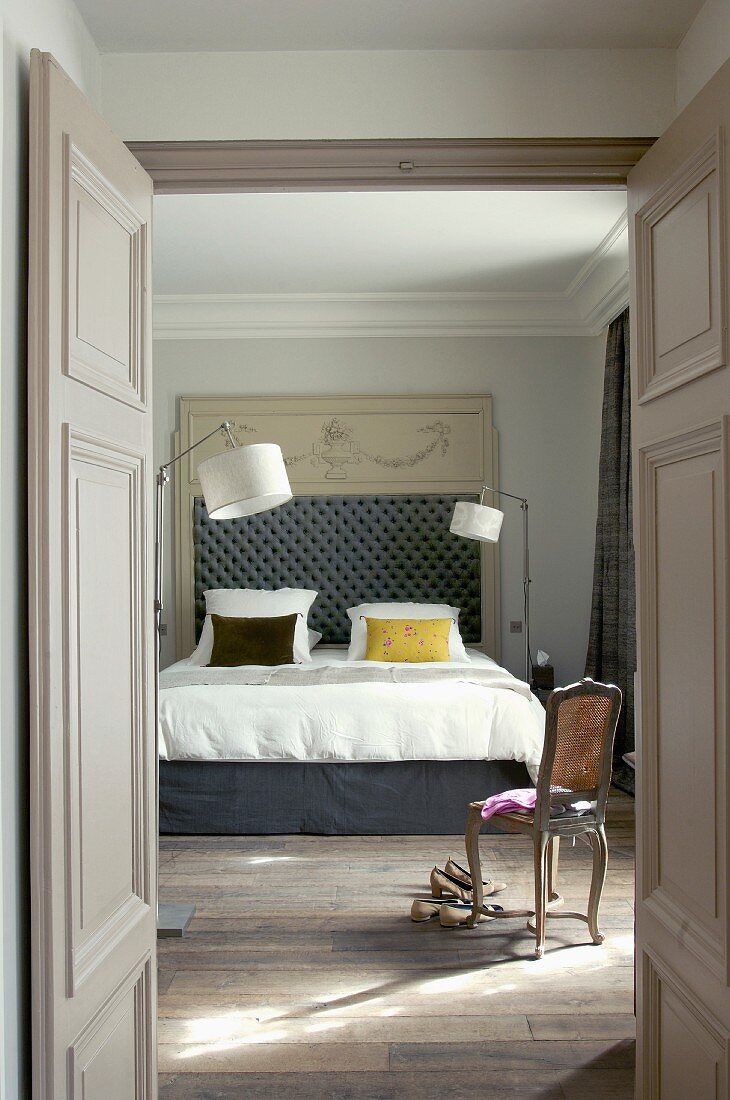 View through open double doors of antique chair in front of double bed with upholstered headboard and retro-style standard lamps