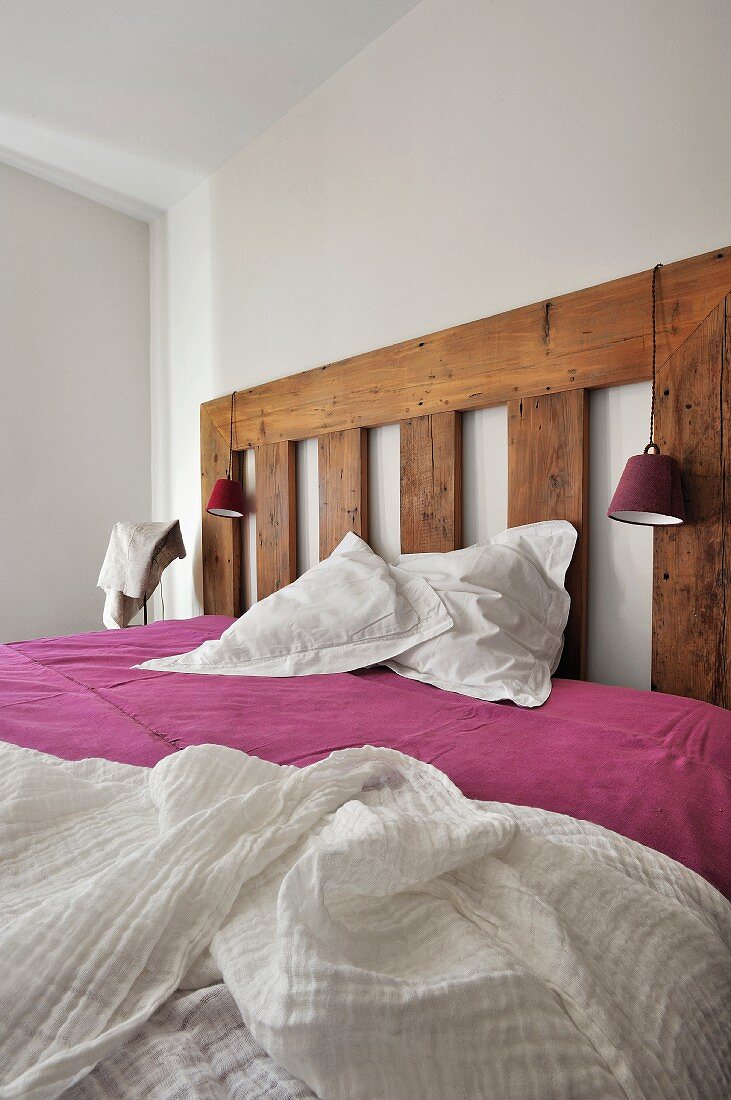 Bett mit Kopfteil aus rustikalem Holz und weisses Betttuch auf violettem Bezug in minimalistischem Schlafzimmer