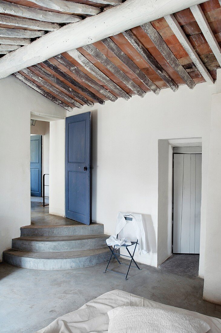 Betonstufen vor blauer Holztür und rustikale, offene Dachuntersicht in puristischem Schlafraum