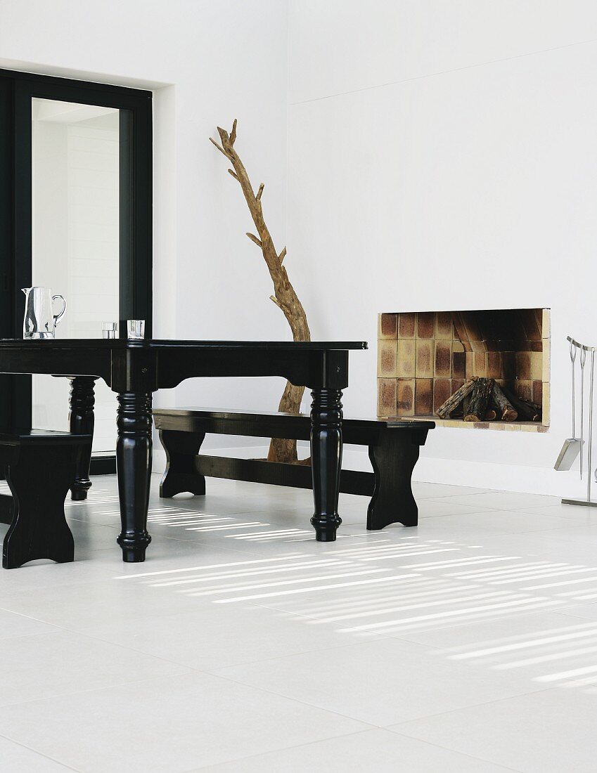 Starke Kontraste in Wohnraum mit schwarz lackiertem Essplatz im Antikstil auf weißem Fliesenboden; offener Ziegelkamin und Baumstammskulptur in der Ecke