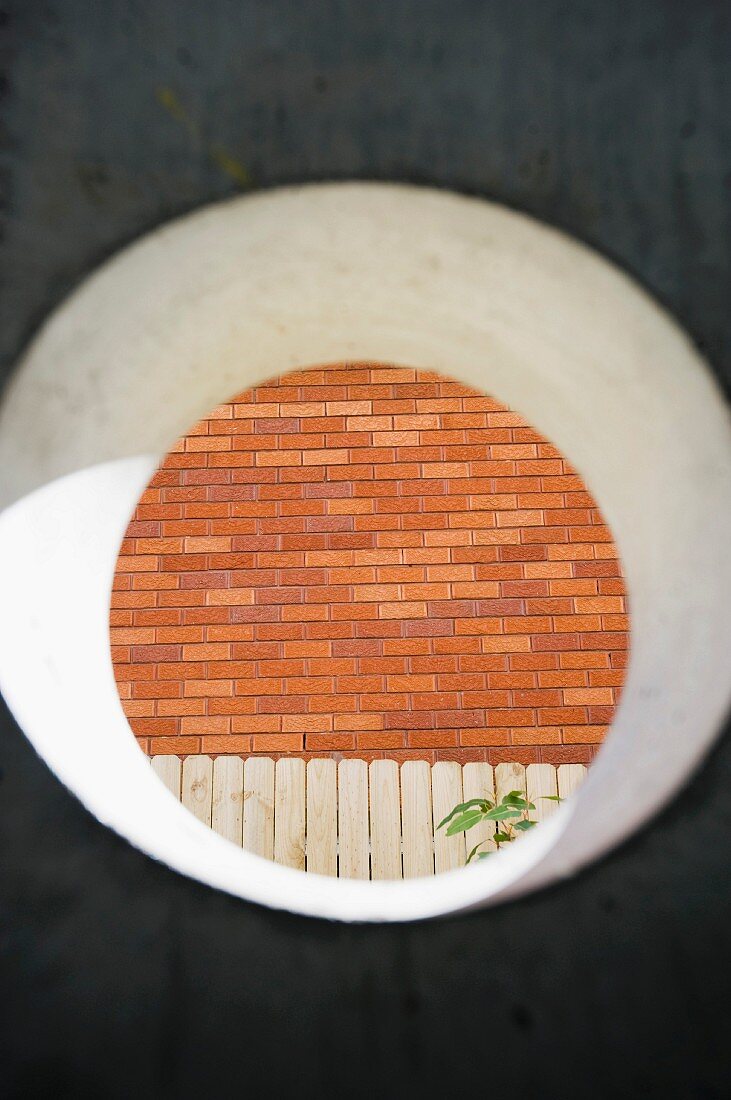 Blick durch kreisförmigen Ausschnitt in Betonwand auf Ziegelfassade und weissen Lattenzaun