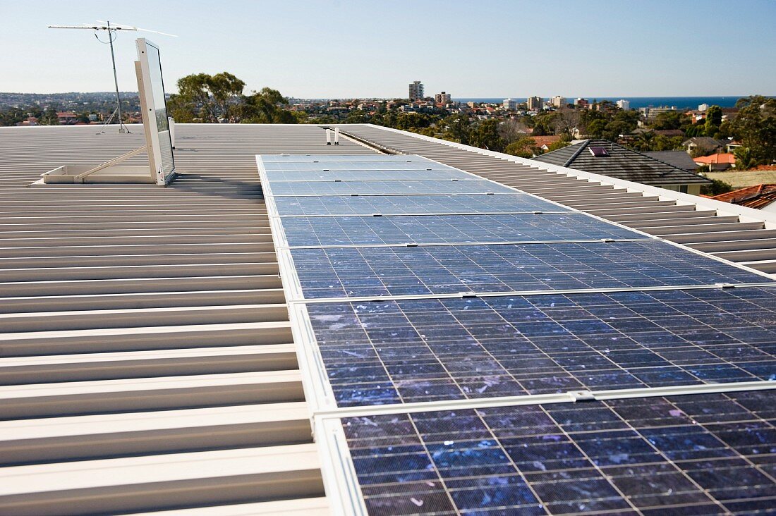 Solar panels on a flat, ribbed roof with open roof hatch; view across the city to the sea