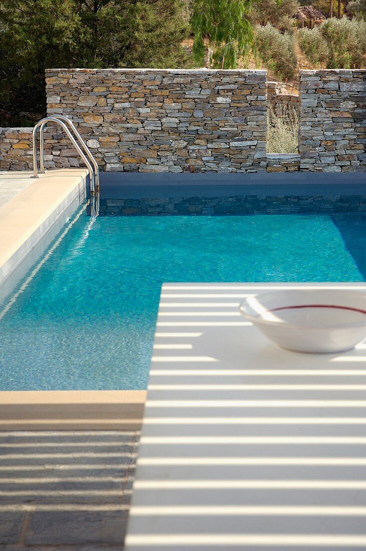 Pool mit Natursteinmauer in mediterraner Landschaft; Licht- und Schattenspiel auf einem Tisch