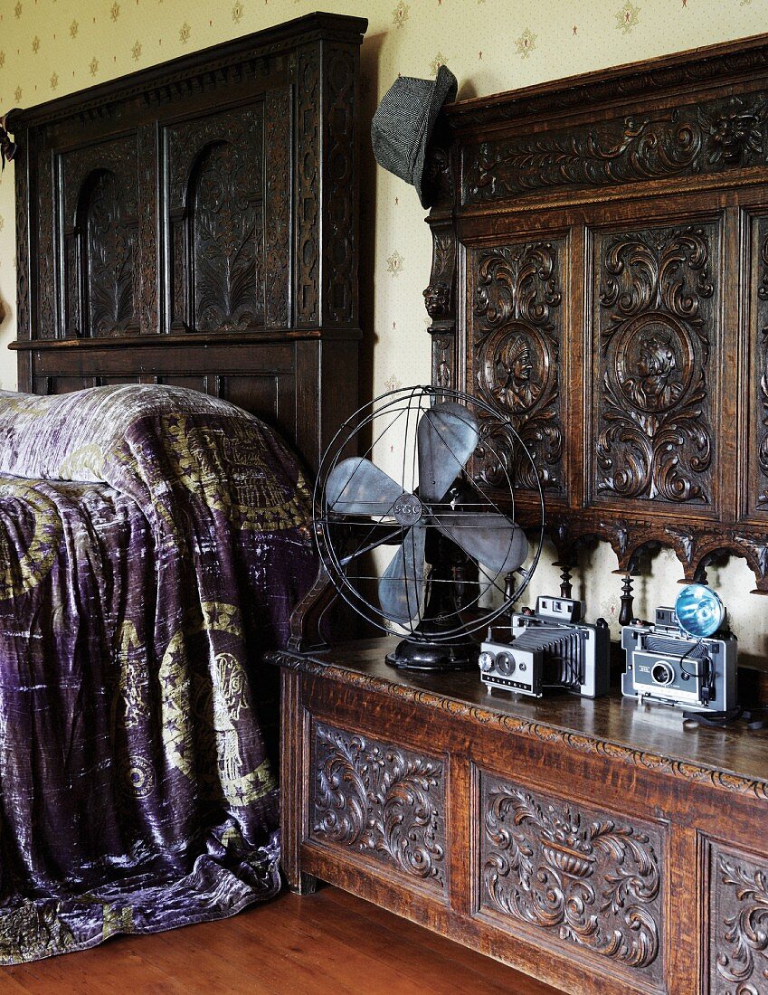 Antiker Ventilator und Kameras auf geschnitzer Holzbank mit hohem Rückenteil neben Bett mit Kopfteil aus dunklem Holz und violett schimmernder Tagesdecke