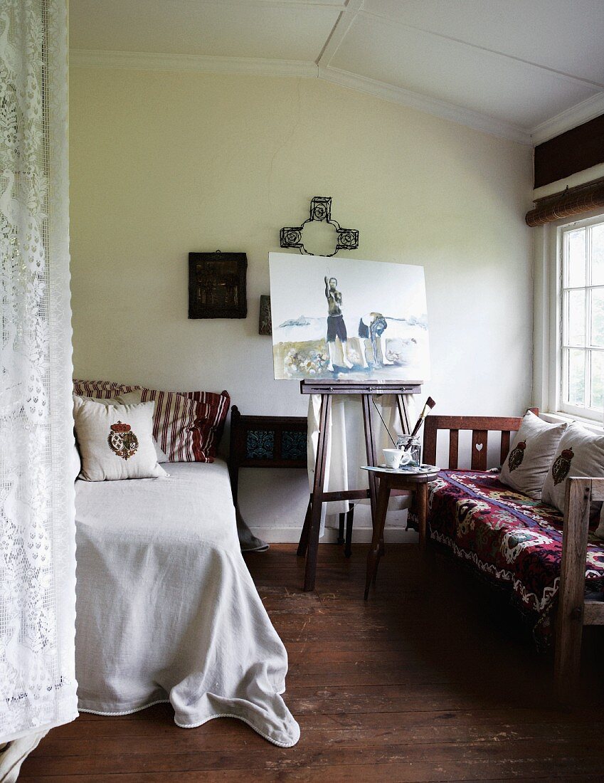 Staffelei mit Bild zwischen Bett und Sitzbank am Fenster, an der Seite teilweise sichtbarer Spitzen Vorhang in schlichtem Schlafzimmer