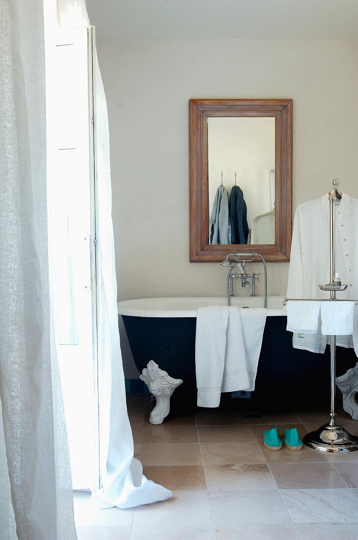 Towels and man's shirts on valet stand in front of vintage clawfoot bathtub below framed mirror on wall in modern interior