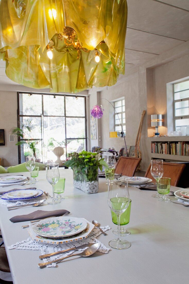Designer-Kronleuchter über gedecktem Tisch mit Blümchentellern und Spitzensets in klassischem Loftraum mit Industriefenstern und Sichtbeton