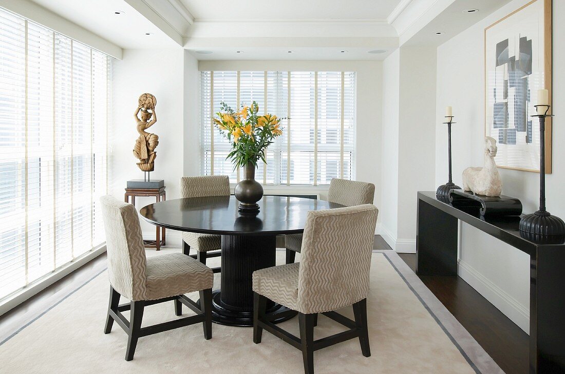Round, dark wood table and upholstered chairs in dining room with floor-to-ceiling windows and half-closed blinds