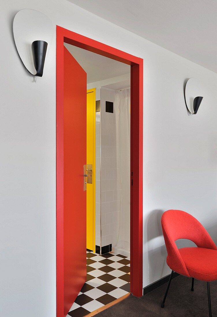 Roter gepolsterter Stuhl an Wand neben offener, roter Tür und Blick in Vorraum mit schwarz-weißem Schachbrettboden