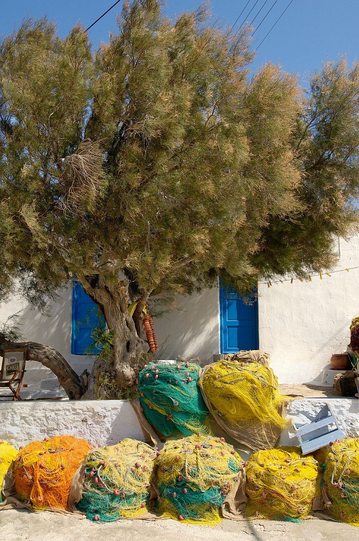 Aufgerollte, farbige Fischernetze vor mediterraner Terrasse und alter Baum vor Haus mit blauen Türläden