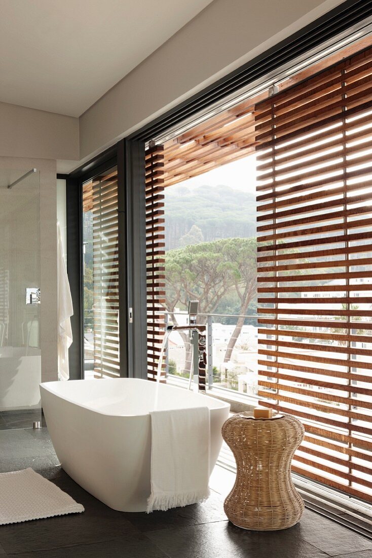 Moderne, freistehende Badewanne und Beistelltisch aus Rattan vor Fensterfront mit teilweise geschossenen Holz Jalousien