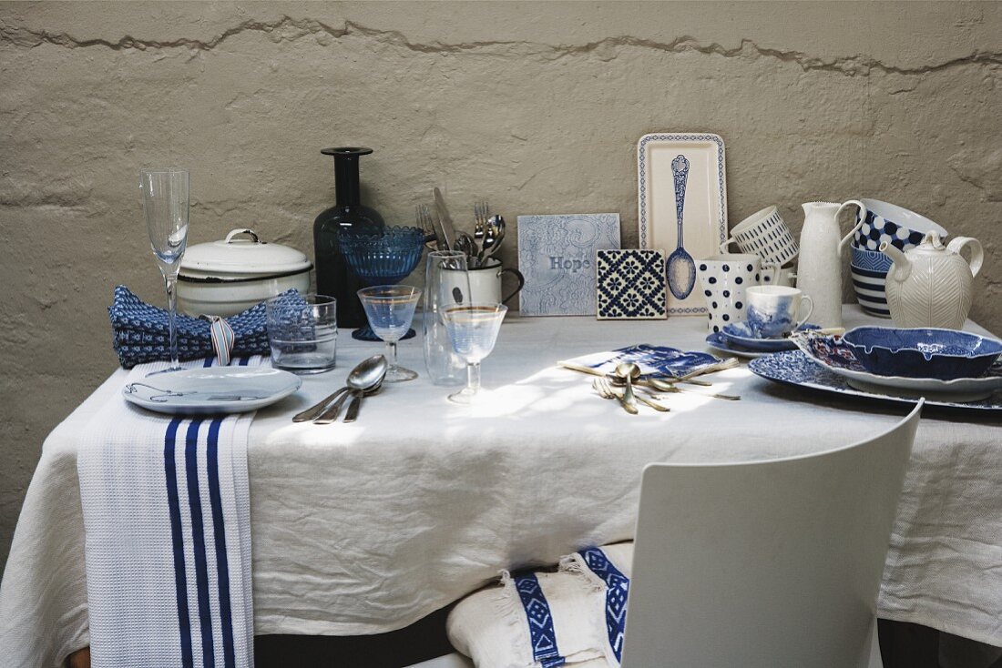 Weiß-blaues Geschirr und Gläser auf weißem Tischtuch und Tisch vor rustikaler verputzter Wand