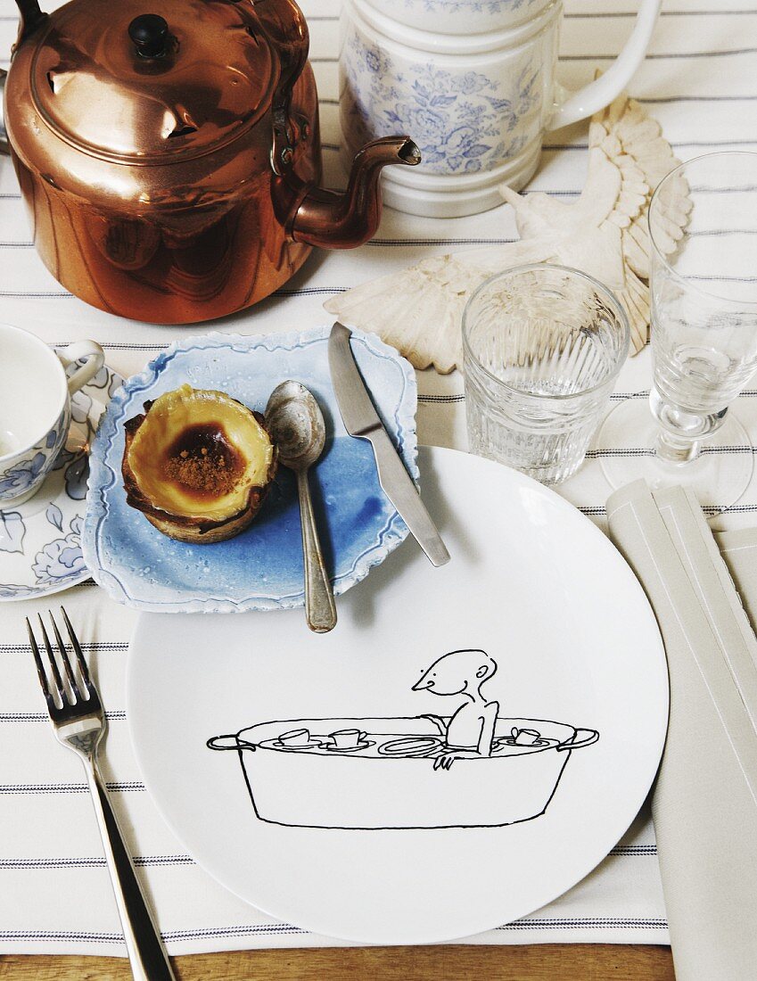 Place setting with comic-style line drawing on plate and ceramic saucer next to copper kettle