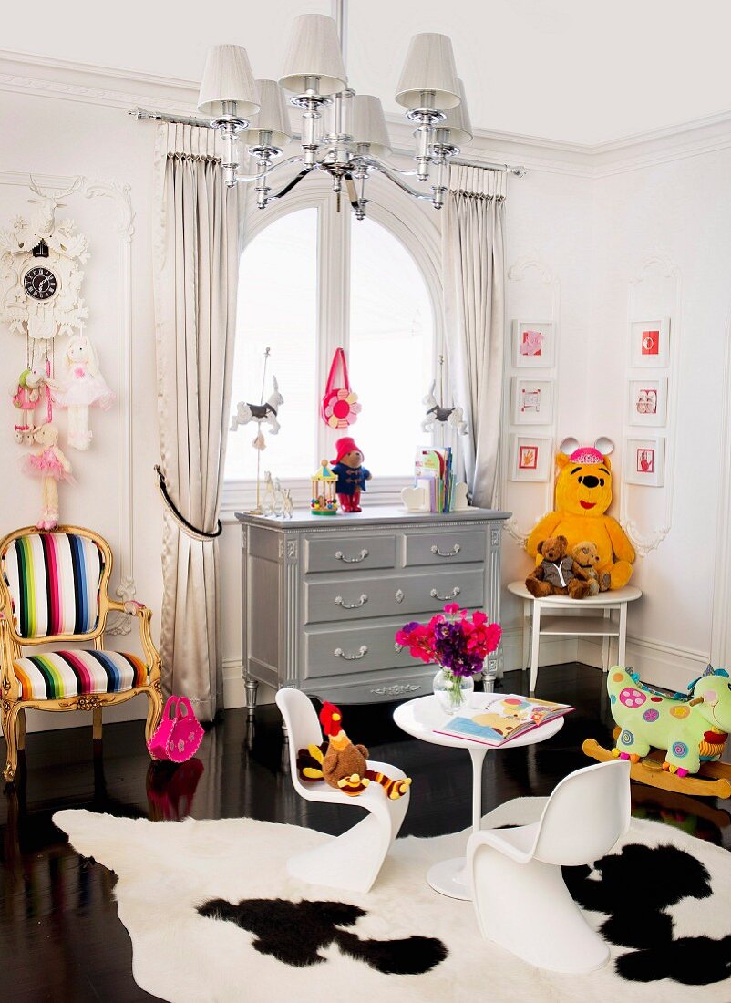 Kinderzimmer in postmodernem Stil, weiße Kunststoff Schalenstühle auf Kuhfell und Neo-Rokoko Sessel neben Kommode am Fenster