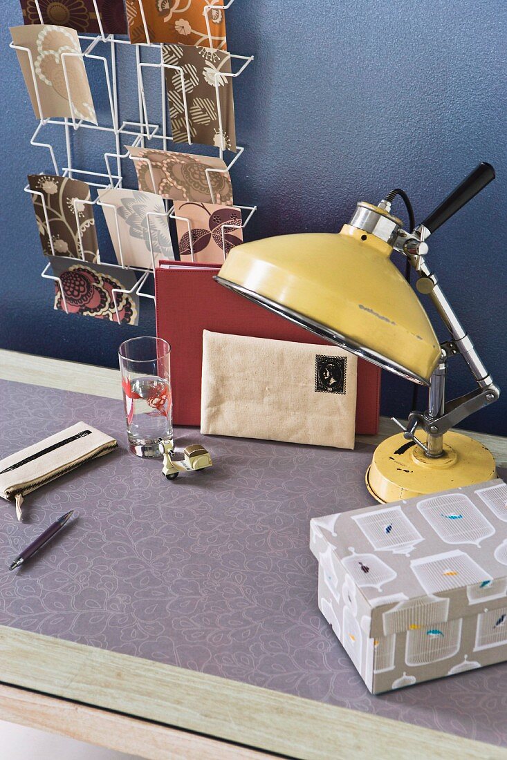 Retro Tischleuchte aus gelb lackiertem Metall und Schreibutensilien auf tapeziertem Tisch vor blauer Wand und aufgehängter Postkartenhalter aus Metall
