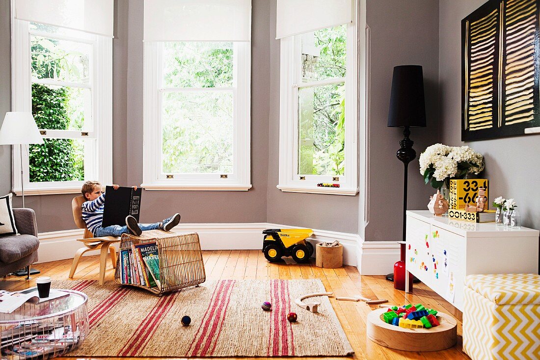 Little boy on a chair with picture book and toys thrown around the room in an elegant bay window with gray walls