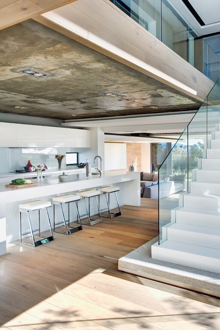 Galley kitchen under exposed concrete ceiling and free standing stairs with glass banister in an open living room