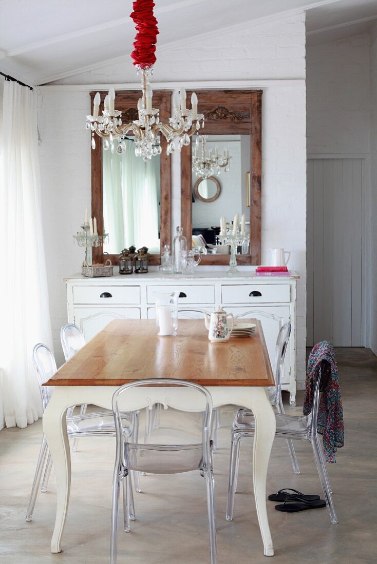 Plastic chairs around a dining table with curved legs and simple wooden top under a chandelier, in front of a chest of drawers and mirror in an open dining area
